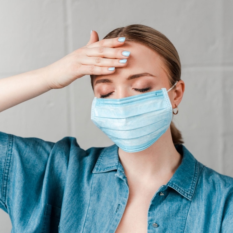 Lady wearing a mask. She has her eyes closed and holds her hand to her forehead. She looks stressed.