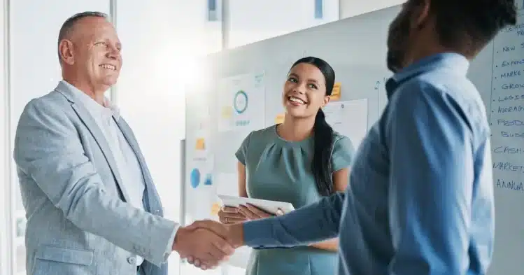 A white man and a black man shake hands and a woman smiles in the background. It's an office environment, after a business agreement.