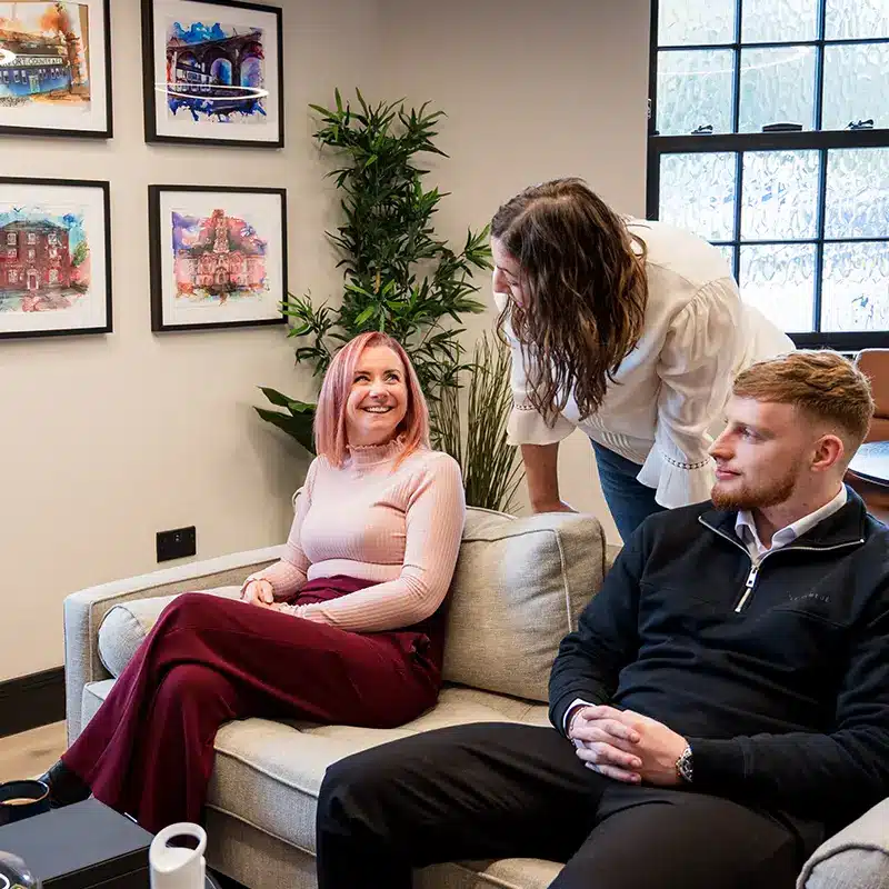Emma, sat on a sofa next to Luke, and Marta, overlooking them behind the sofa, discuss a marketing campaign for Adria Solutions
