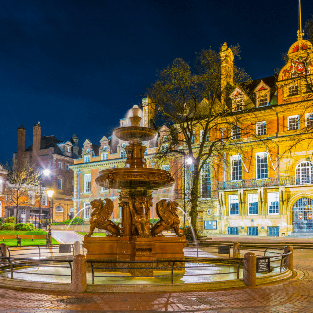 Illuminated fountain in Town Hall Square, Leicester, surrounded by historic buildings at night.