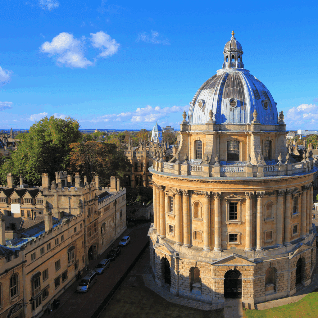 Aerial view of the Radcliffe Camera, an iconic domed building in Oxford, England, surrounded by historic university architecture under a bright blue sky.