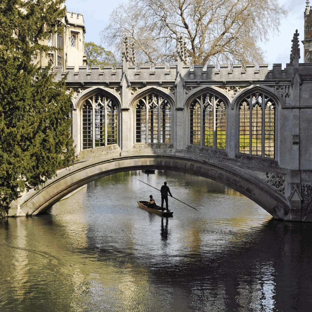 A small punt glides under the ornate stone arch of the Bridge of Sighs at St John's College, Cambridge, framed by gothic windows and tranquil waters.
