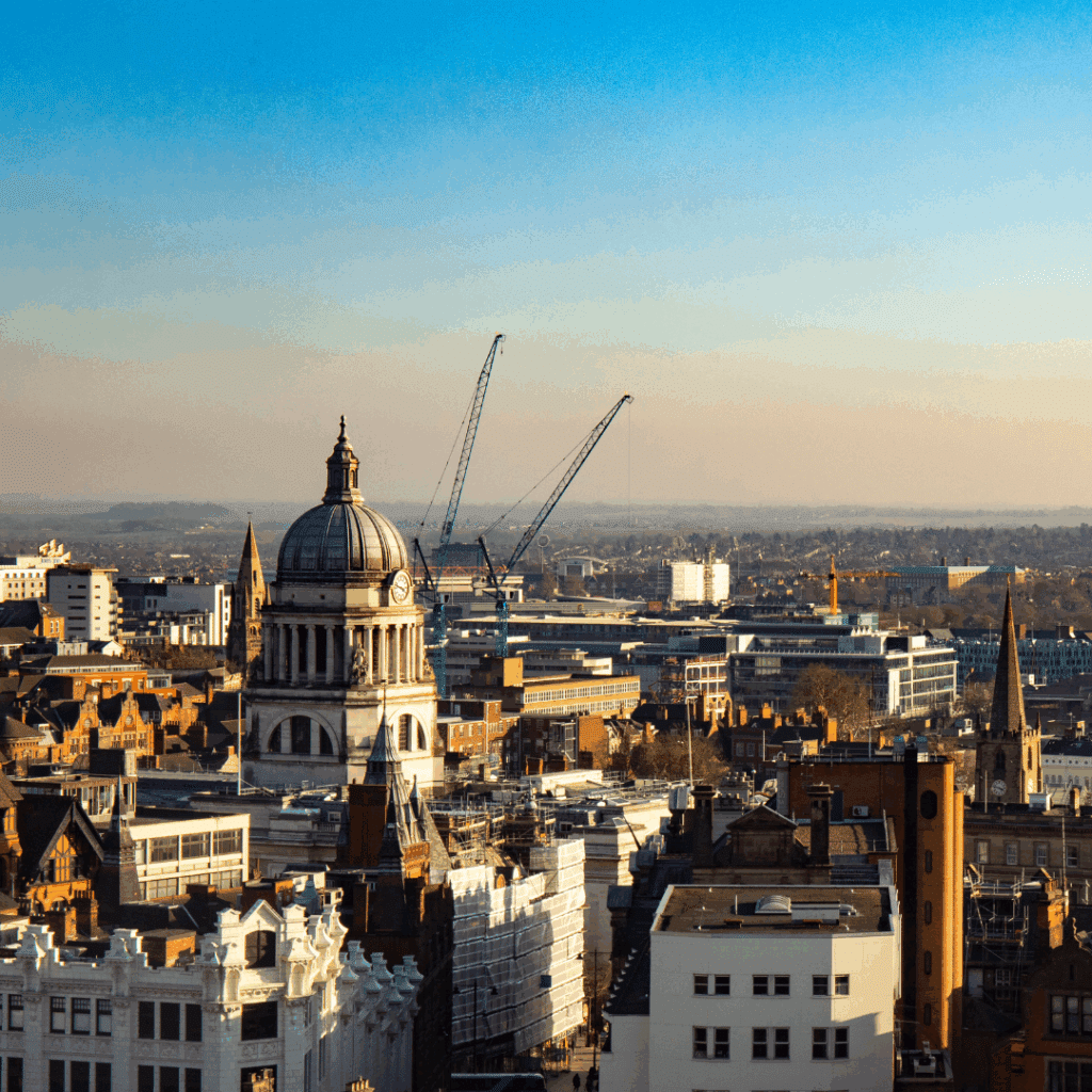 A panoramic cityscape of Nottingham at sunset, with cranes and the Council House dome visible among a mix of old and new buildings.