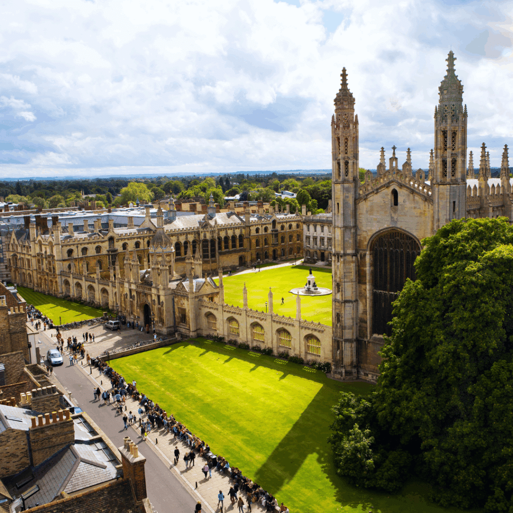 A wide-angle aerial view of King's College Chapel and the surrounding college buildings in Cambridge, with tourists walking below and neatly manicured lawns.