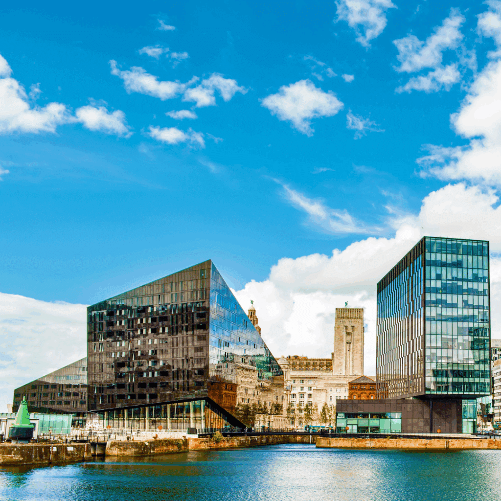 Contemporary glass buildings reflecting the Liverpool skyline across the water, with a clear blue sky overhead.