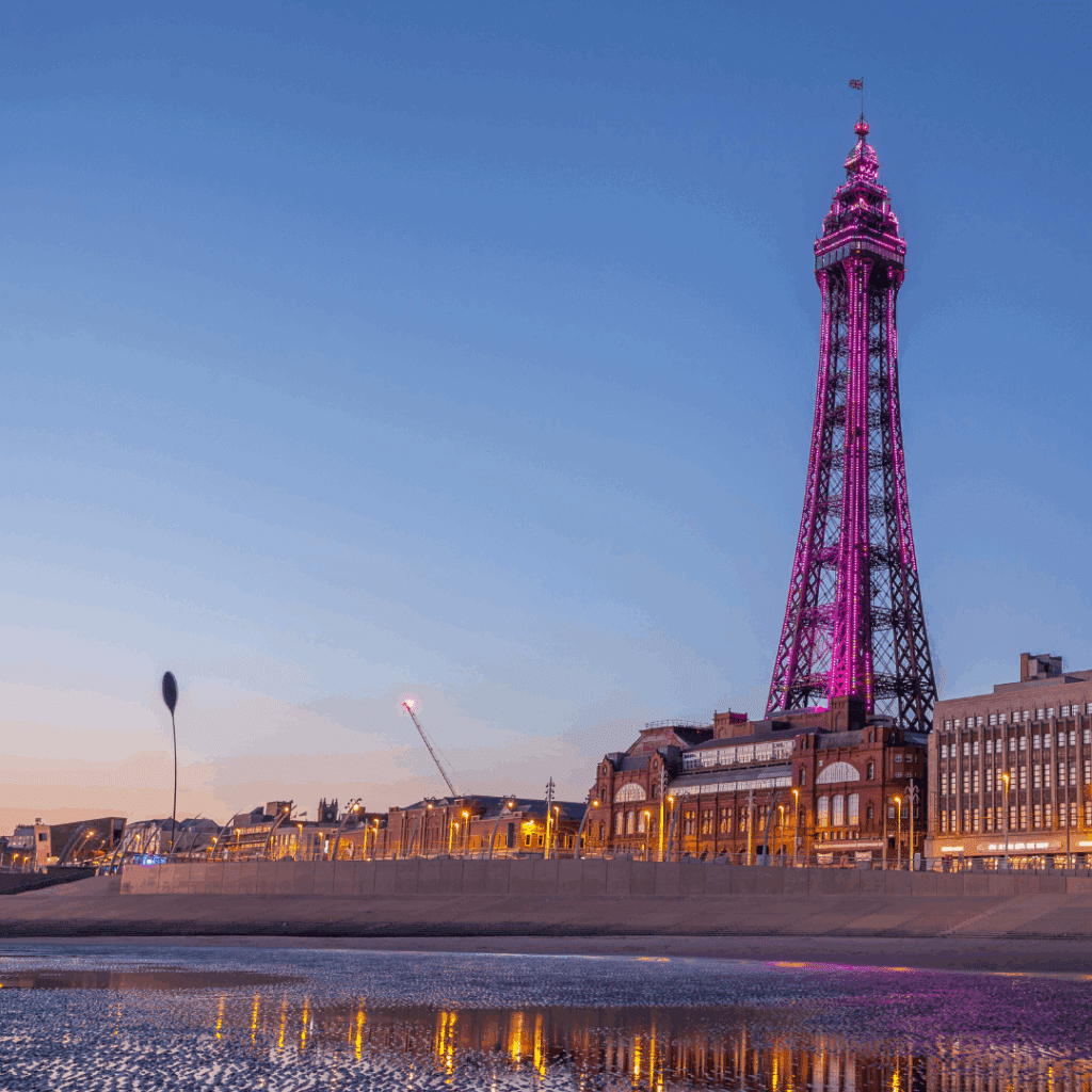 The iconic Blackpool Tower glowing pink at dusk on the Lancashire coastline, reflecting in the water below.