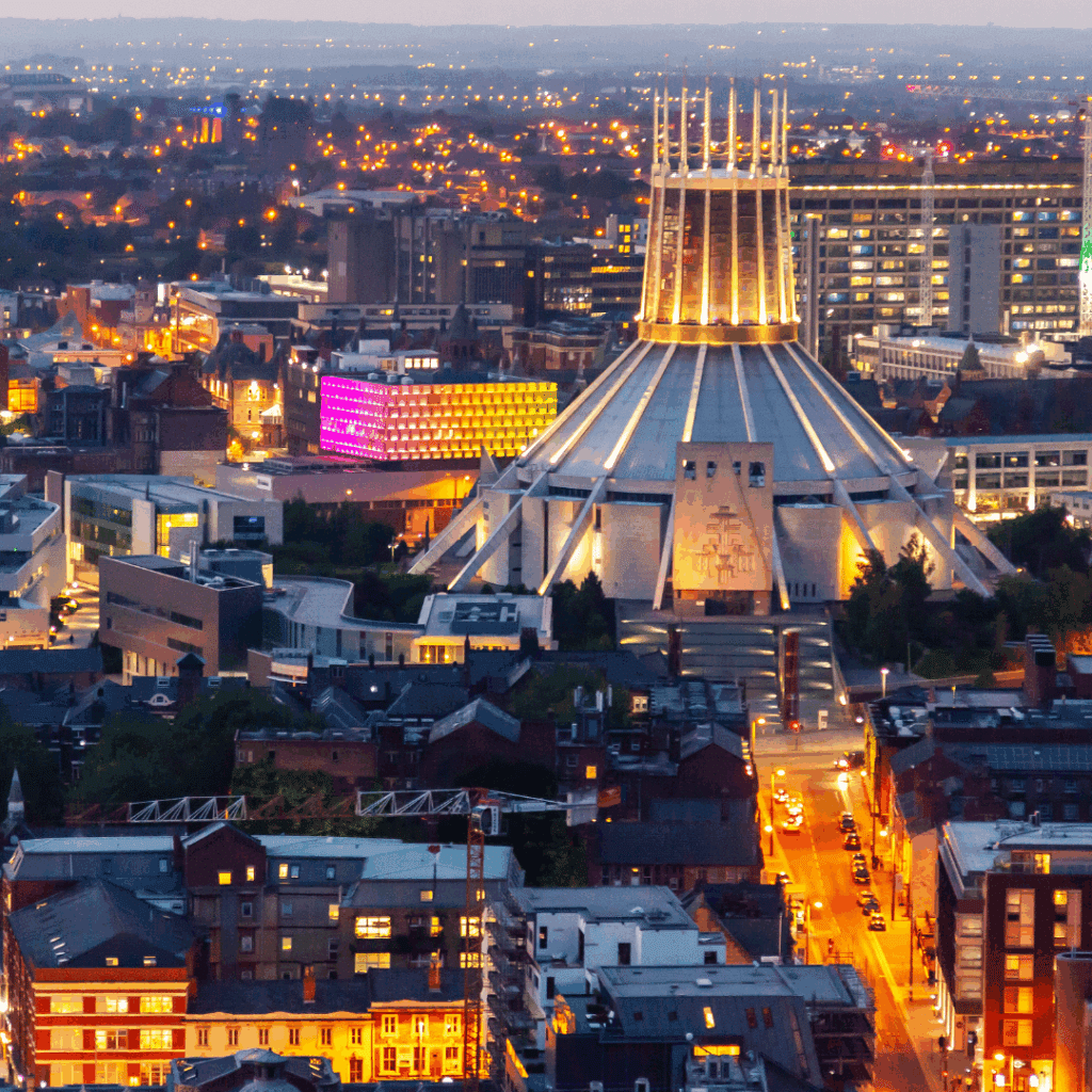 Aerial view of Liverpool Metropolitan Cathedral lit up at night, surrounded by glowing city lights and colourful buildings.