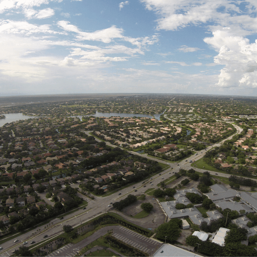 Aerial view of a sprawling suburban neighbourhood with houses, roads, and small lakes under a partly cloudy sky.