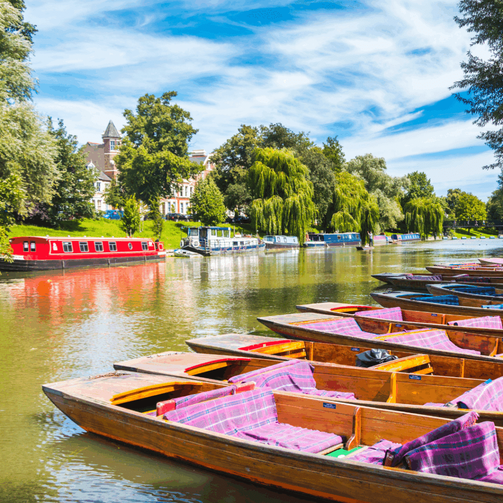 A row of colorful wooden punts lined along the bank of the River Cam in Cambridge, with narrowboats and lush greenery in the background.