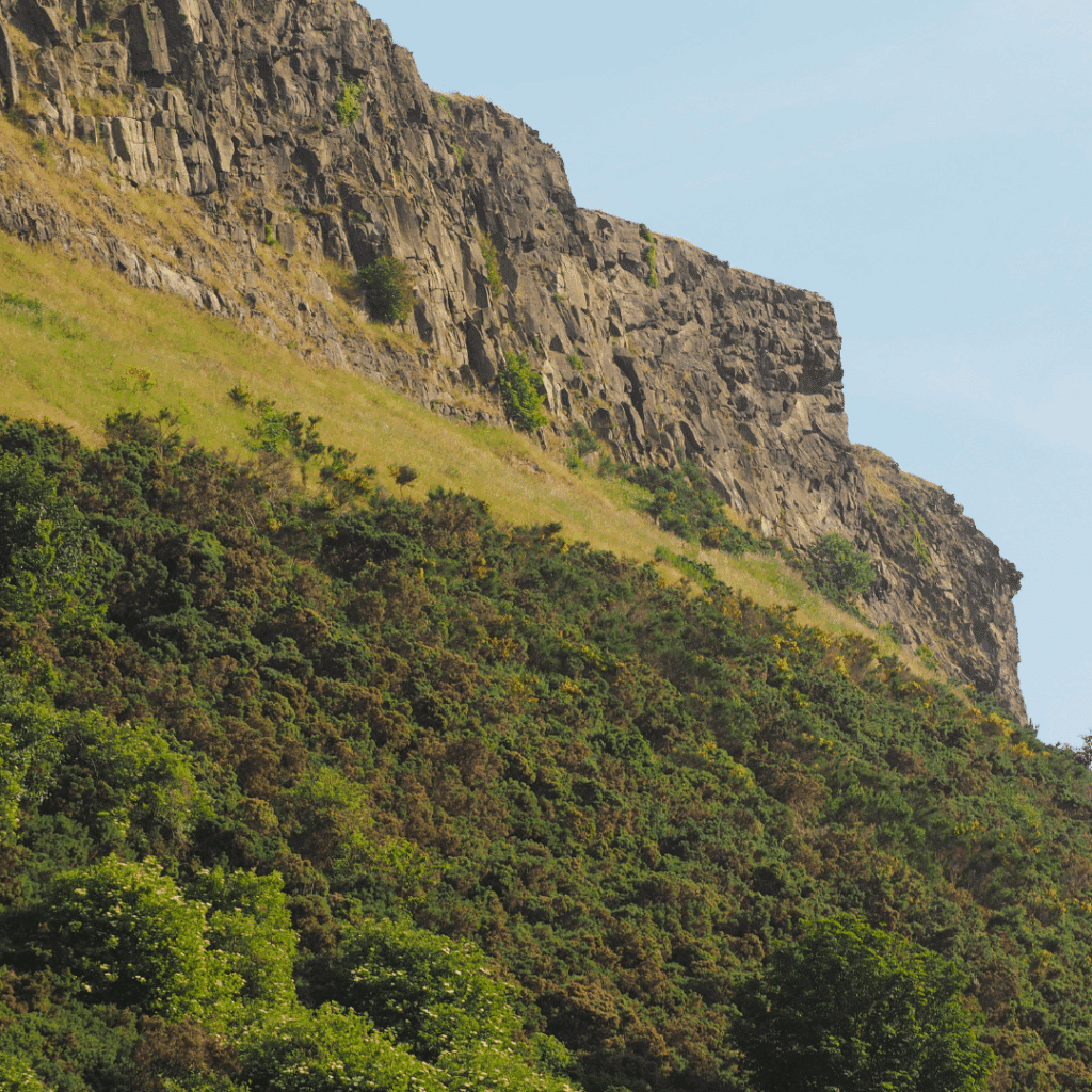 Natural rocky outcrop of Arthur’s Seat covered with grass and shrubs, under blue skies.