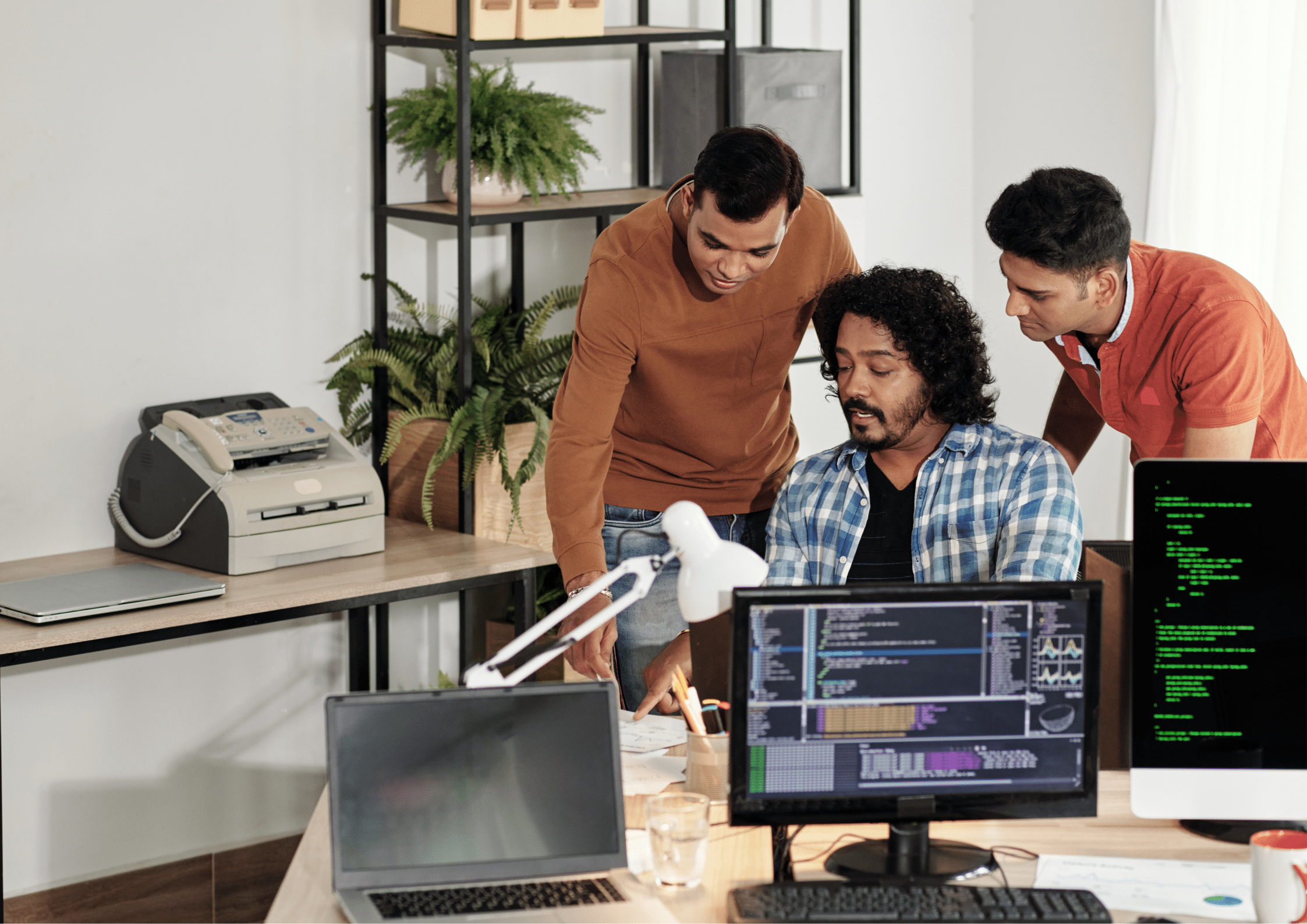 Three male developers working together around a desk with laptops and computer monitors displaying code in a tech office.