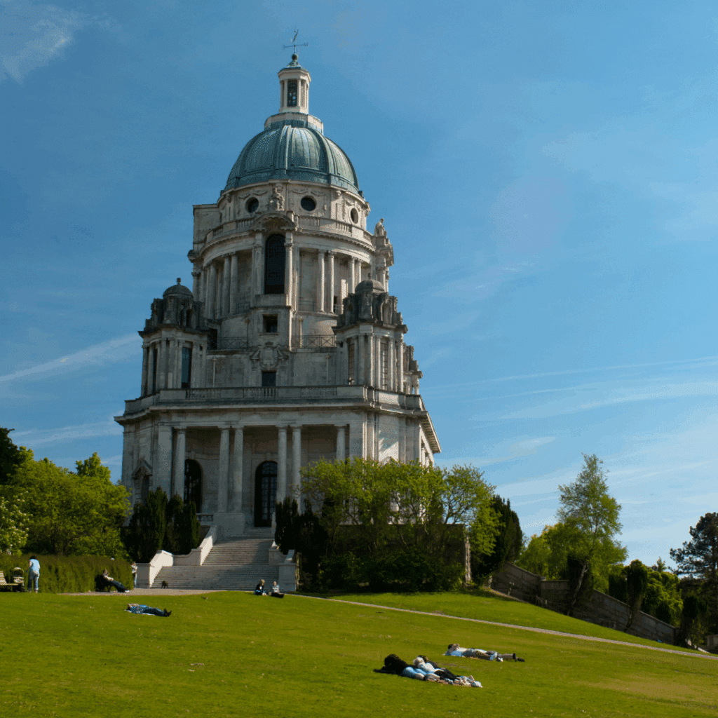 The domed Ashton Memorial in Williamson Park, Lancaster, Lancashire, standing proudly under a blue sky with visitors relaxing nearby.
