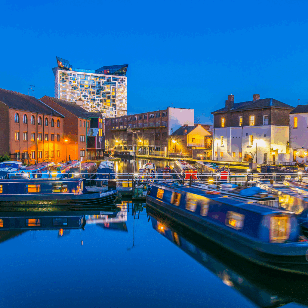 Night view of Birmingham's canal network with moored boats and The Cube building illuminated in the background.