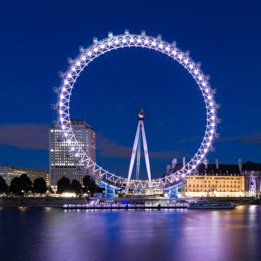 Night view of the London Eye lit up with purple lights, reflecting off the River Thames