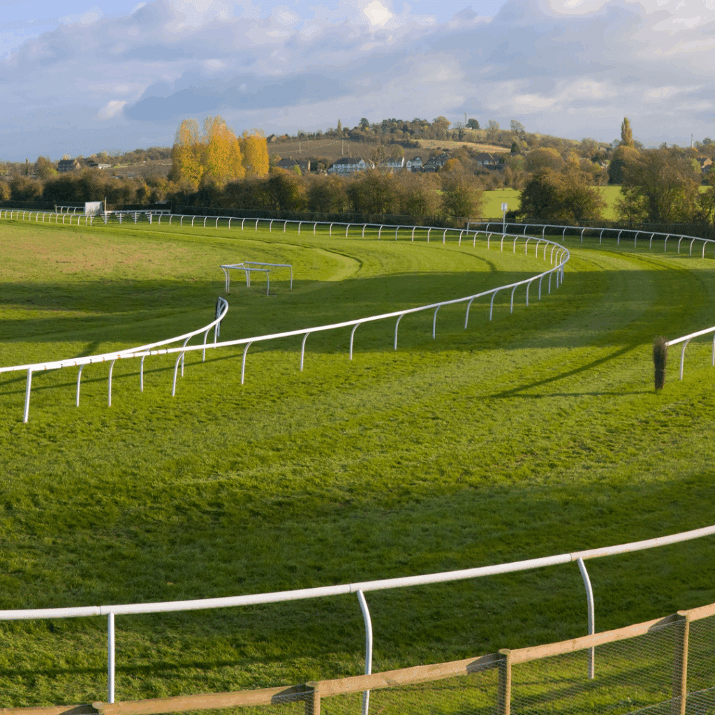 Curved, fenced horse racing track in a lush green rural landscape under a partly cloudy sky.