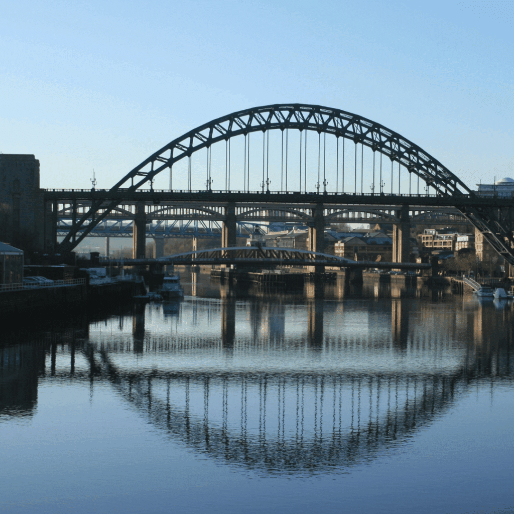 Multiple bridges spanning the River Tyne in Newcastle, including the iconic Tyne Bridge, with clear water reflections on a quiet morning.