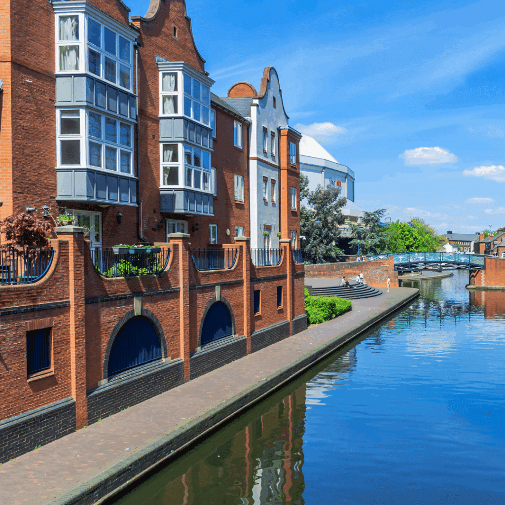 A peaceful canal scene in Birmingham with red-brick buildings and footbridges under a clear blue sky.