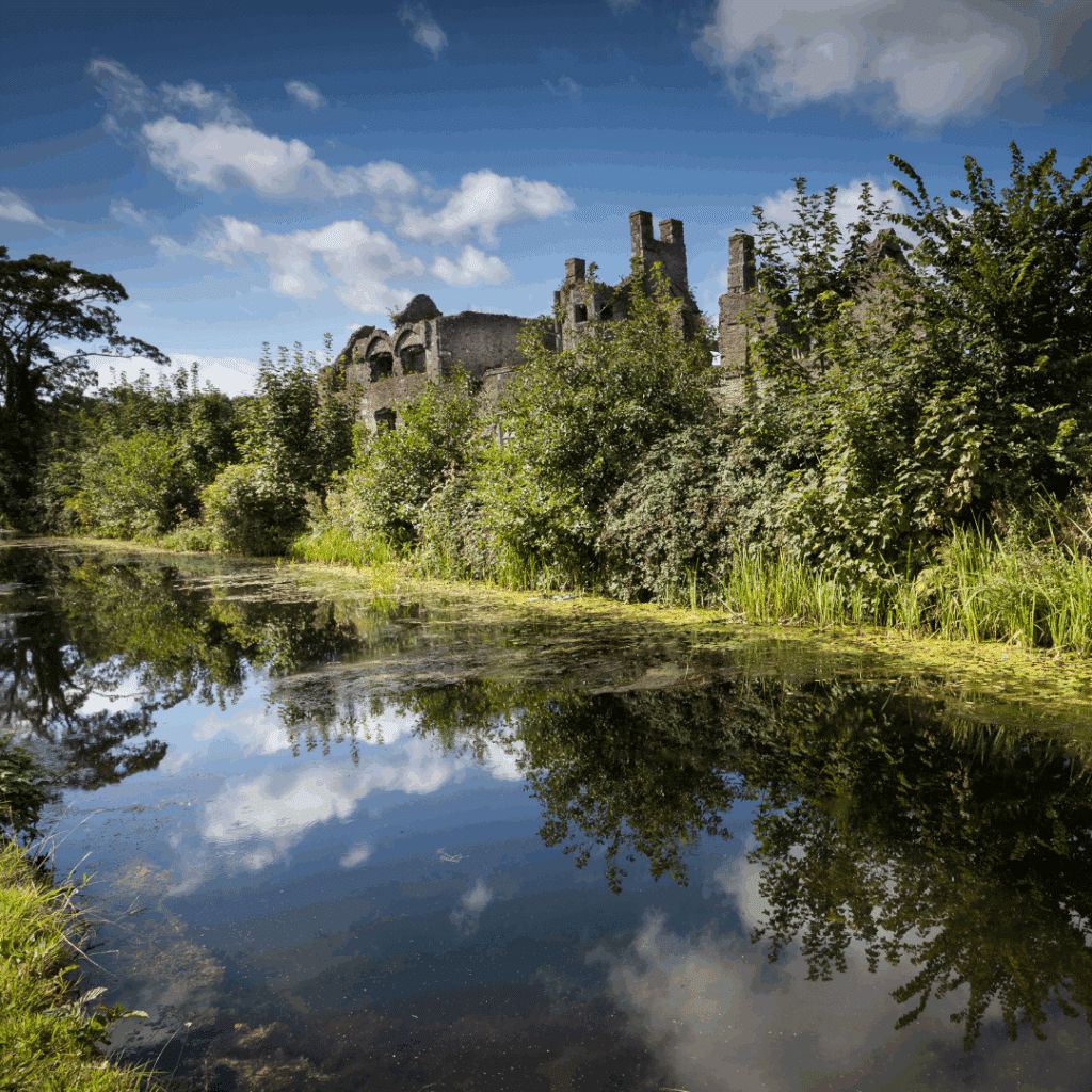 Overgrown ruins of a stone building reflected in a calm canal surrounded by dense greenery and blue sky.