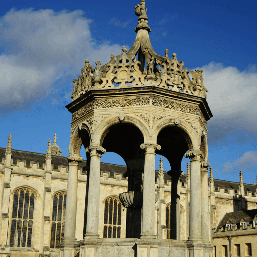 The ornate stone fountain at the center of Trinity College’s Great Court in Cambridge, with gothic buildings and blue sky in the background.