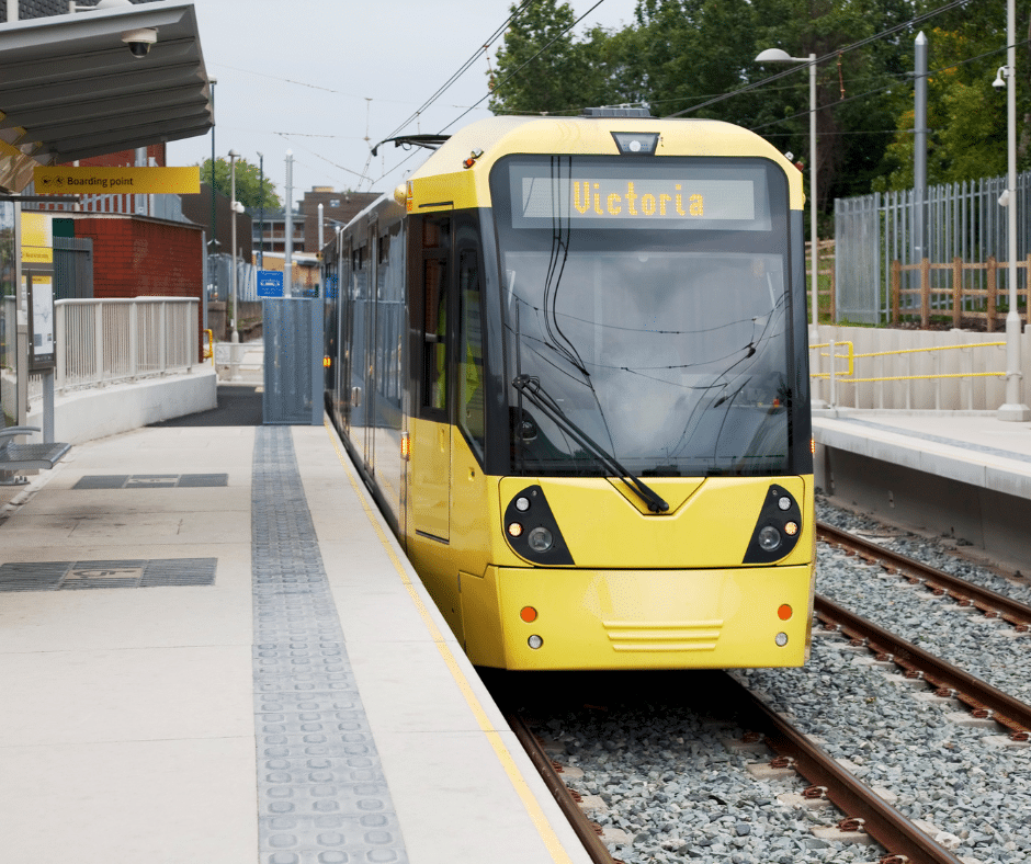 Yellow Manchester Metrolink tram waiting at a modern, open-air platform with a sign showing ‘Victoria’.