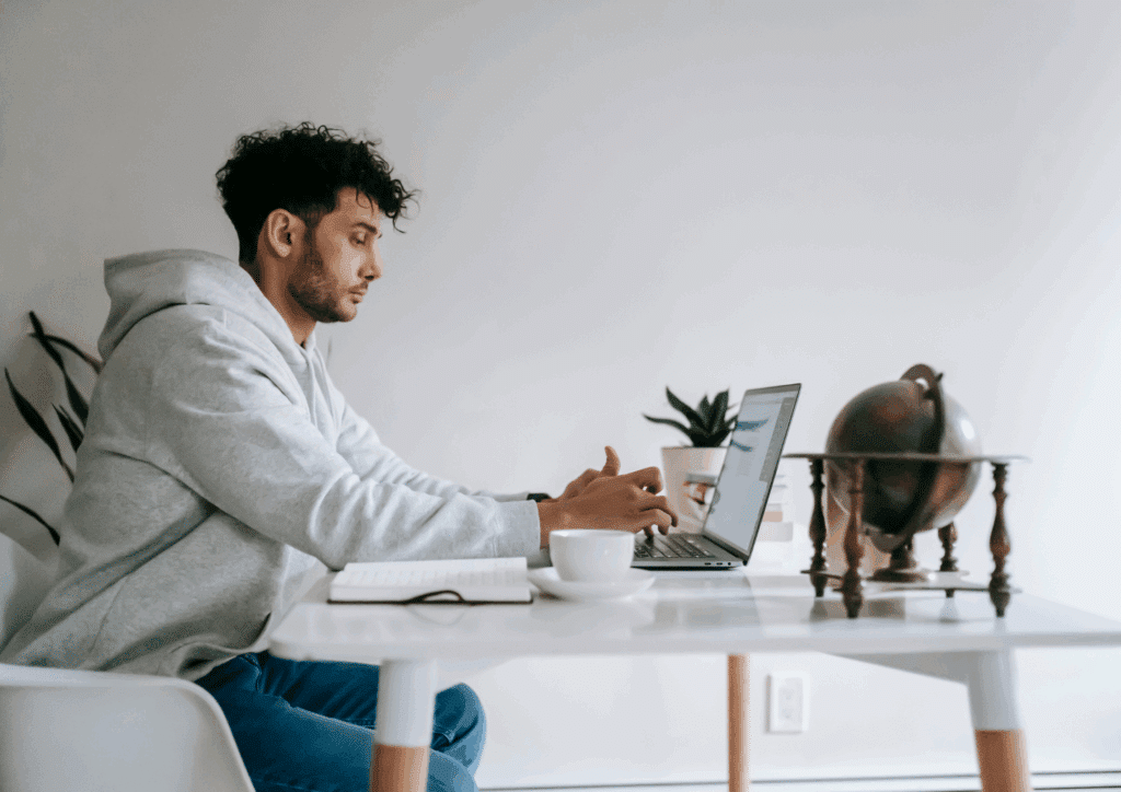 A man wearing a hoodie working on a laptop at a home office desk with a cup of coffee and globe decor.