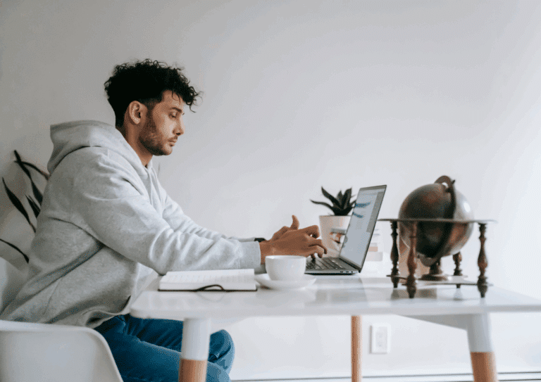 A man wearing a hoodie working on a laptop at a home office desk with a cup of coffee and globe decor.