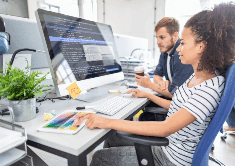 Two coworkers working together at a desk, with HTML/CSS code displayed on a large monitor and a color palette on a tablet, indicating a web development project.