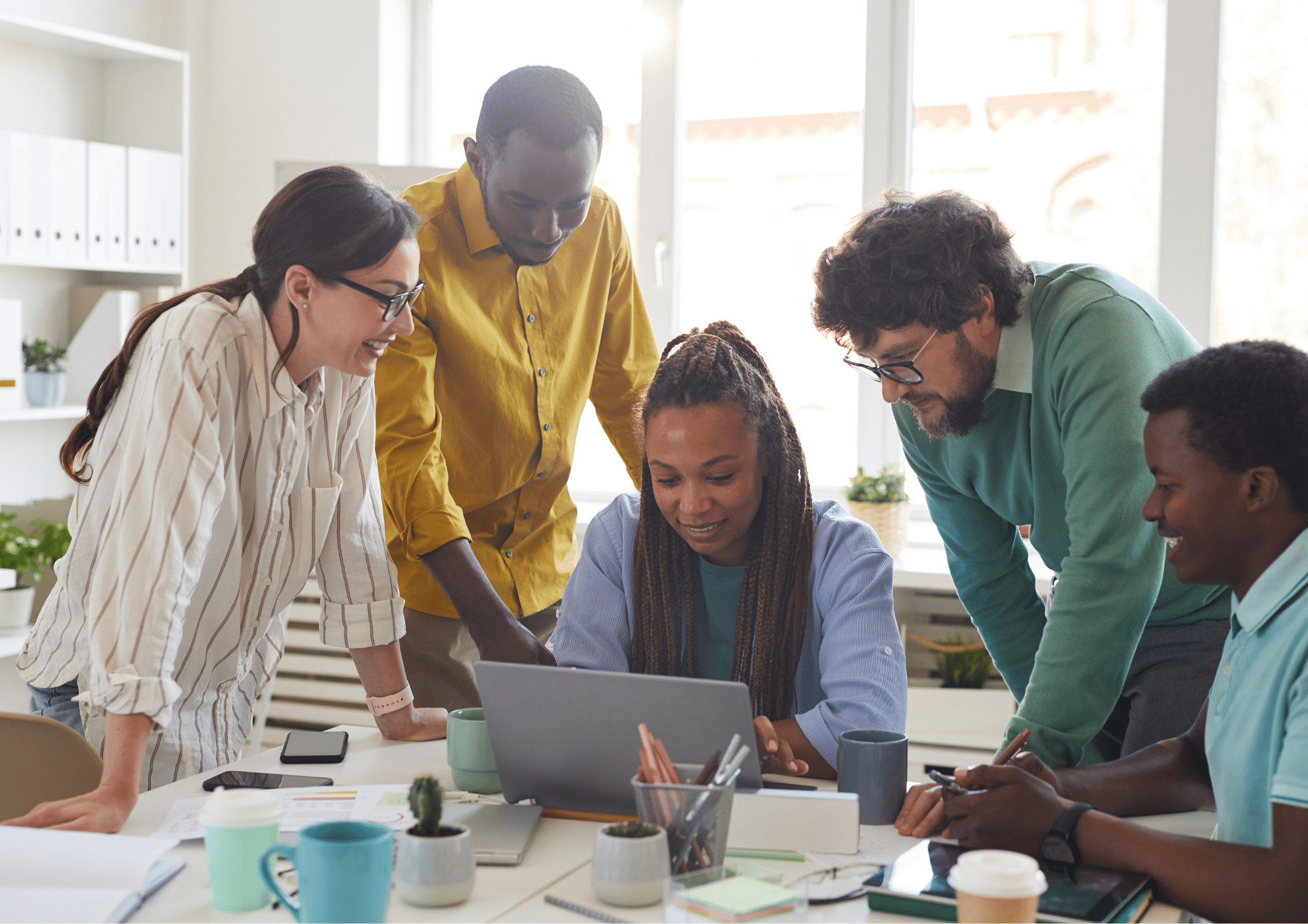 A group of five colleagues gathered around a laptop, smiling and engaged in discussion, in a bright, open office space