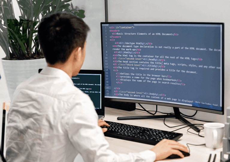 A man in a white shirt working at a desk with dual monitors displaying HTML code.