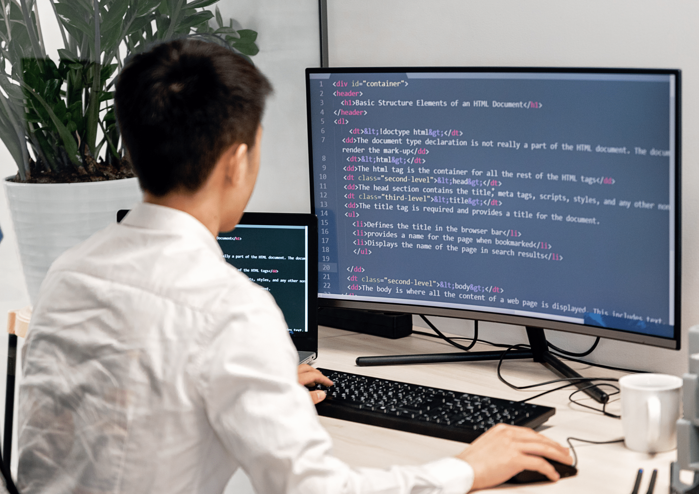 A man in a white shirt working at a desk with dual monitors displaying HTML code.