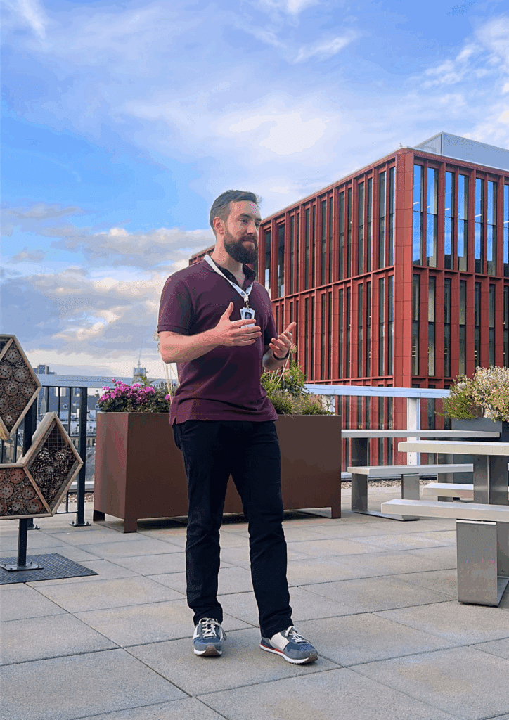 A man delivering a talk on a rooftop terrace with the red façade of a modern office building and a blue sky in the background.