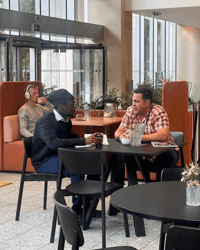 Two men sitting at a round café table in a modern, open atrium, engaged in a focused discussion over coffee and notes.