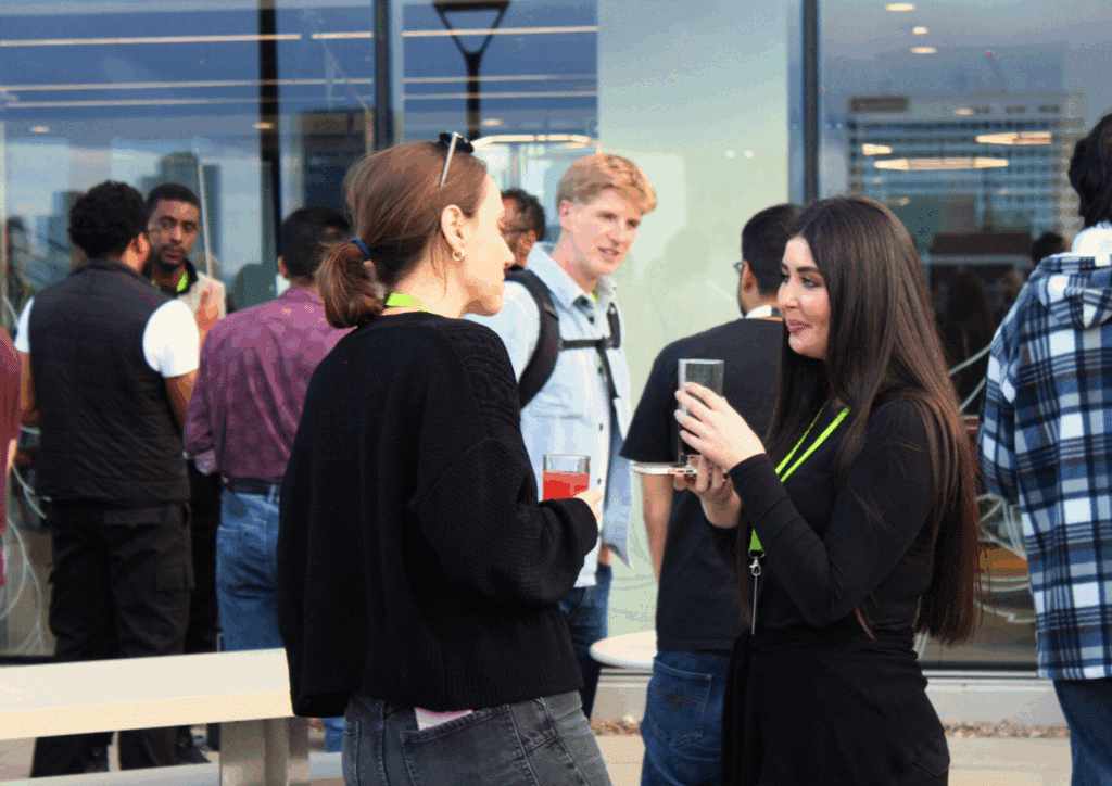 Two women engaged in lively conversation at an outdoor rooftop event, each holding a drink with other guests mingling nearby.