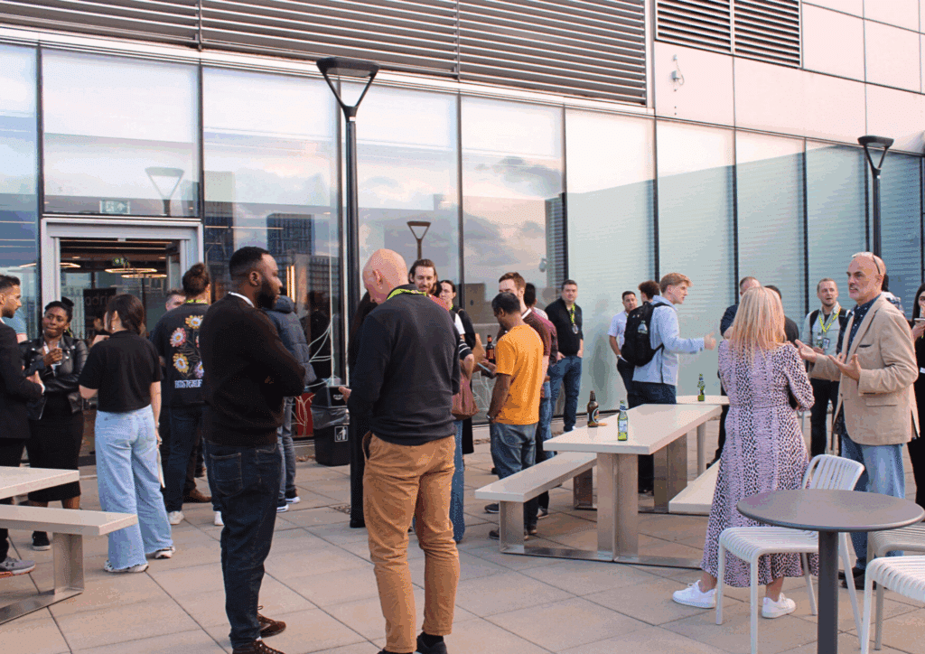 A bustling rooftop gathering with groups of people chatting and networking outside a modern glass building.