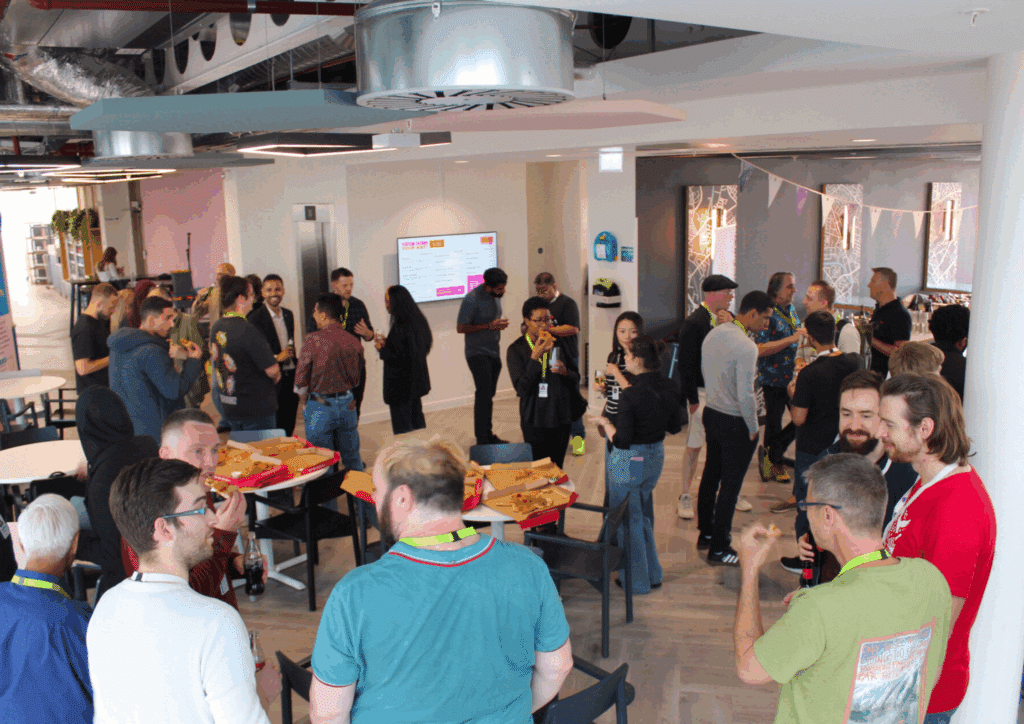 An indoor social gathering with pizza boxes and drinks on tables, where attendees are enjoying food and networking.