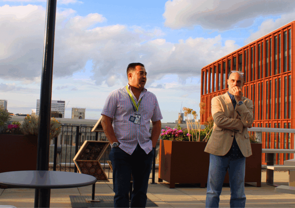 Two men giving closing remarks on a rooftop terrace, set against Manchester’s city skyline in the evening light.
