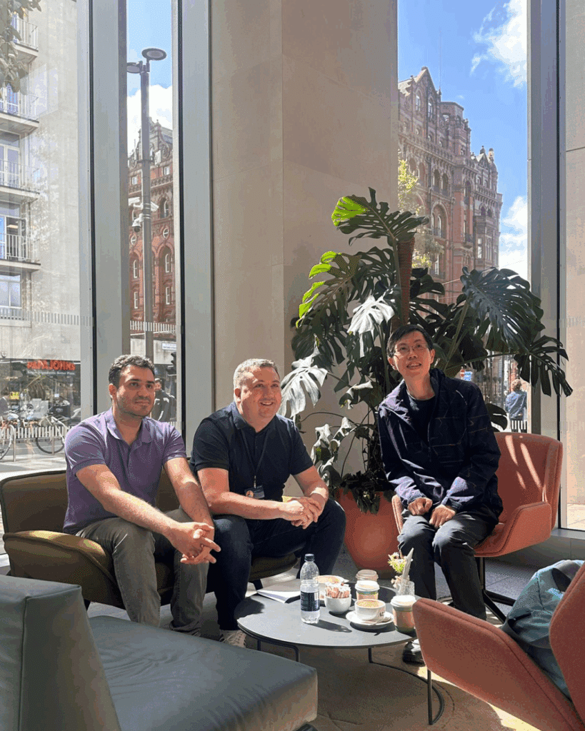 Three men sitting together in a bright, glass-walled lounge, smiling and enjoying a relaxed conversation with drinks on a low table.