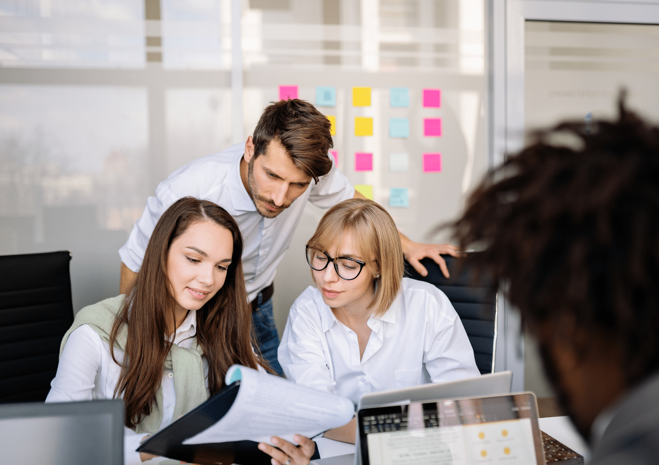 A group of young professionals gathered around a desk reviewing documents and discussing ideas, with laptops and colourful sticky notes on the glass wall in the background.