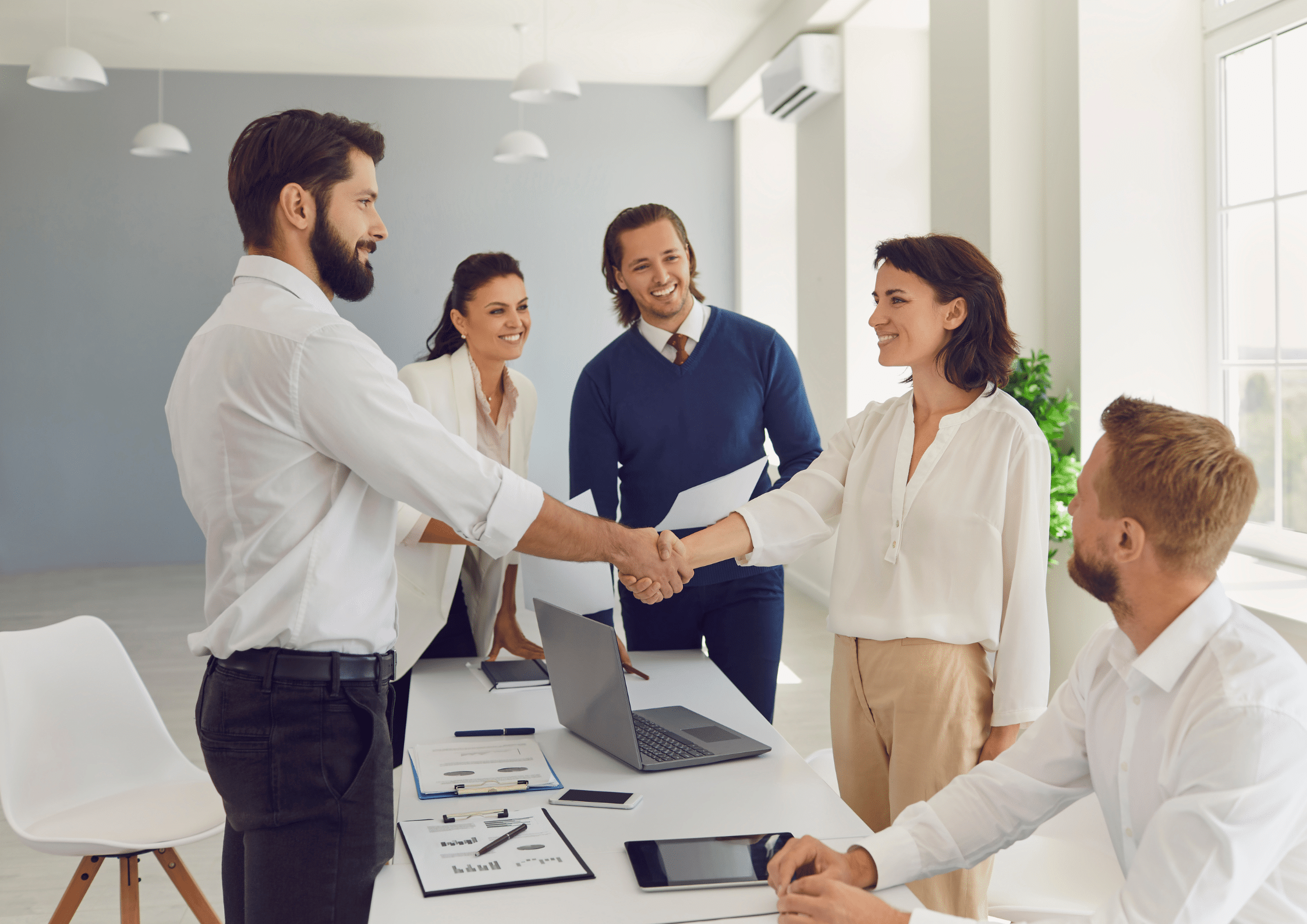 A man and woman shaking hands across a meeting table while colleagues look on, celebrating a business agreement in a bright, modern office setting.