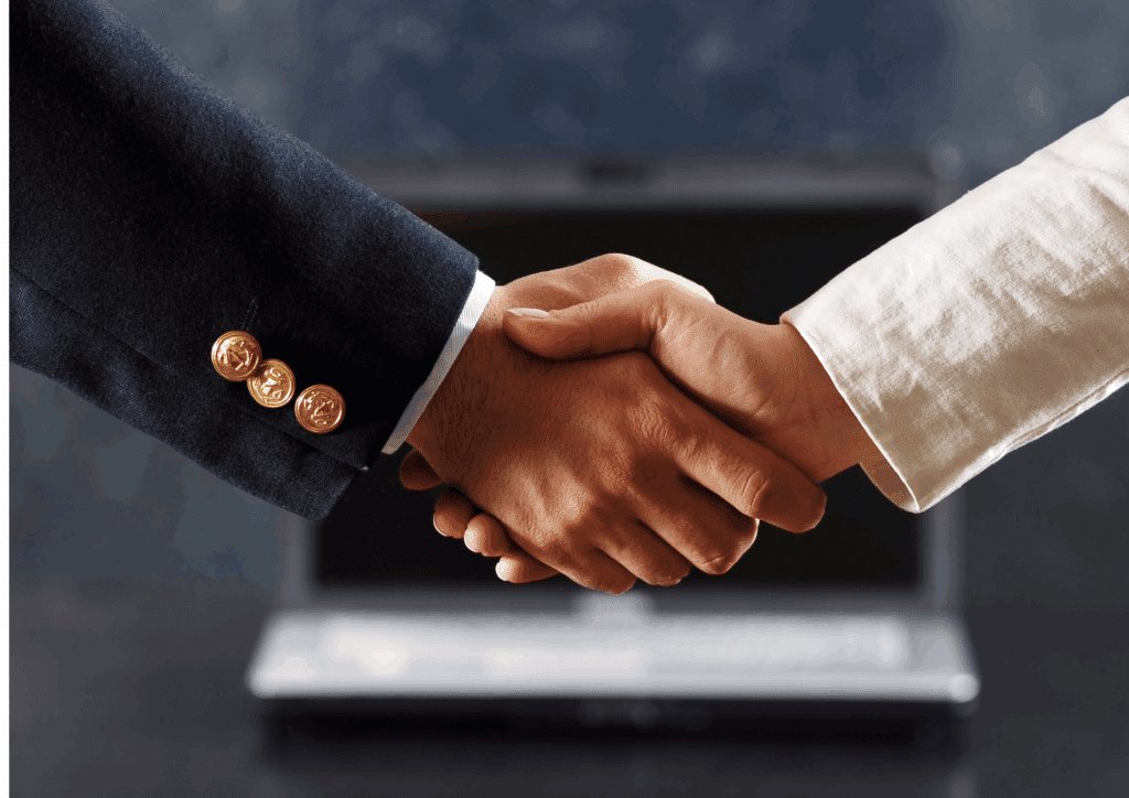 Close-up of a handshake between two professionals, one in a dark suit and the other in a light jacket, with a laptop in the background.