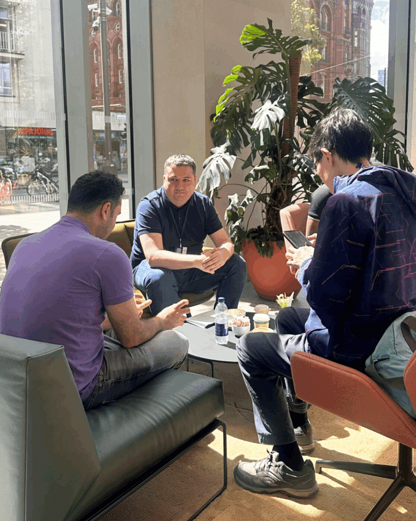 A small group gathered in a sunlit seating area, actively engaged in a conversation with coffee cups and notebooks in view.