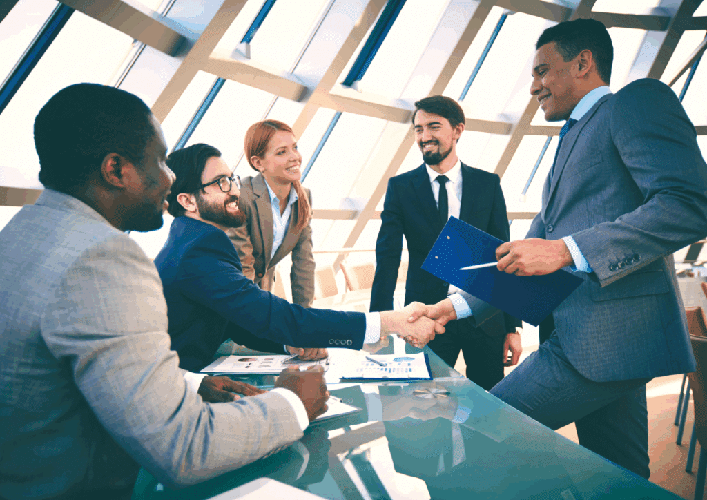 A group of five professionals in business attire gathered around a glass table, smiling and shaking hands in a modern office setting.