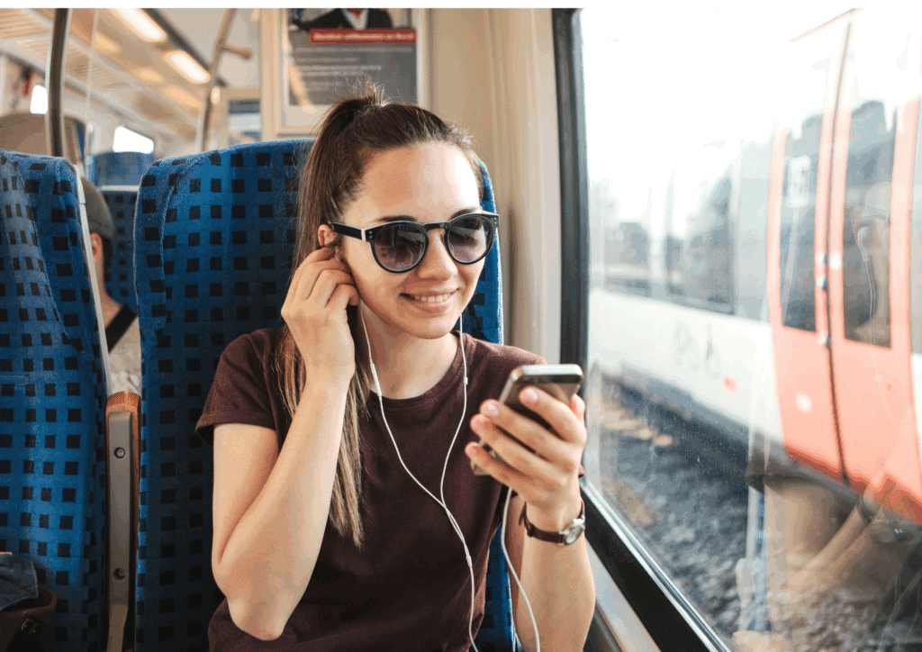A young woman wearing sunglasses listening to music on her phone with earphones while riding a train. She is smiling and looking out the window.