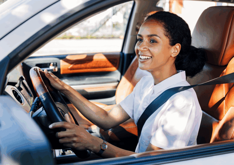 A cheerful woman behind the wheel of a car, wearing a seatbelt and a light pink shirt, enjoying her drive in daylight.