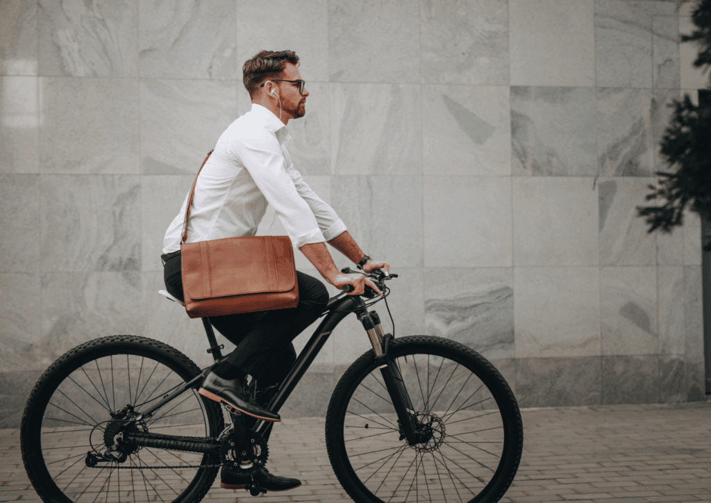 A man dressed in business attire, including a white shirt and brown shoulder bag, riding a black bicycle on a city street.