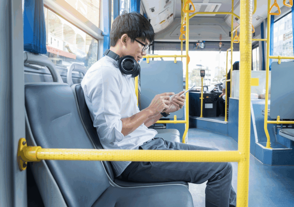 A young man sitting on a bus, wearing glasses and headphones around his neck, engaged with his phone while commuting.
