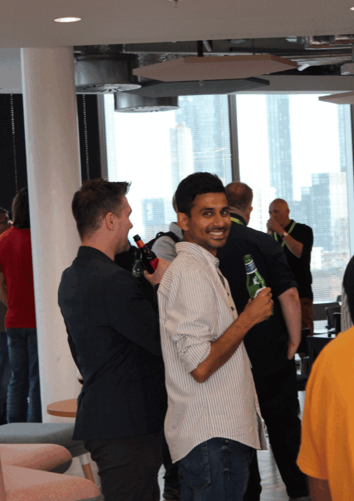A smiling man holding a drink looks toward the camera at a professional social event with others chatting in the background.