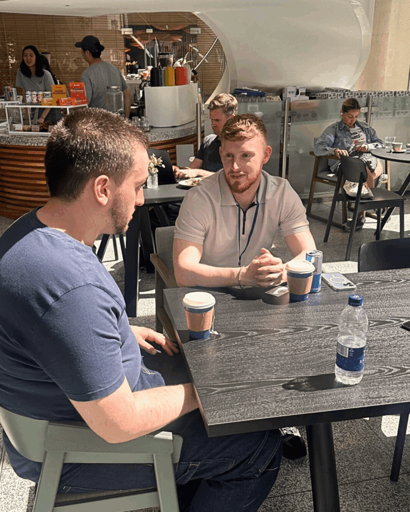 Two colleagues in a well-lit café setting discussing work, with coffee cups, a water bottle, and tech devices on the table.