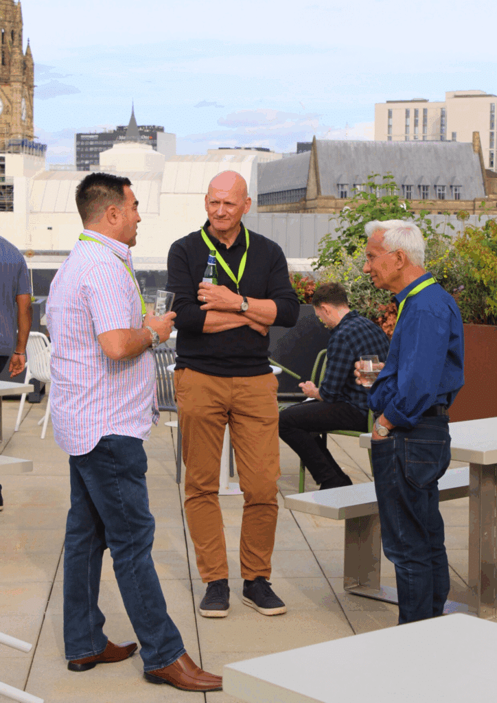 Three men having a relaxed discussion on a rooftop terrace with views of Manchester’s skyline in the background.