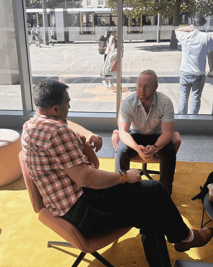Two men seated near a large window, deep in conversation, as natural light pours into the indoor lounge space.